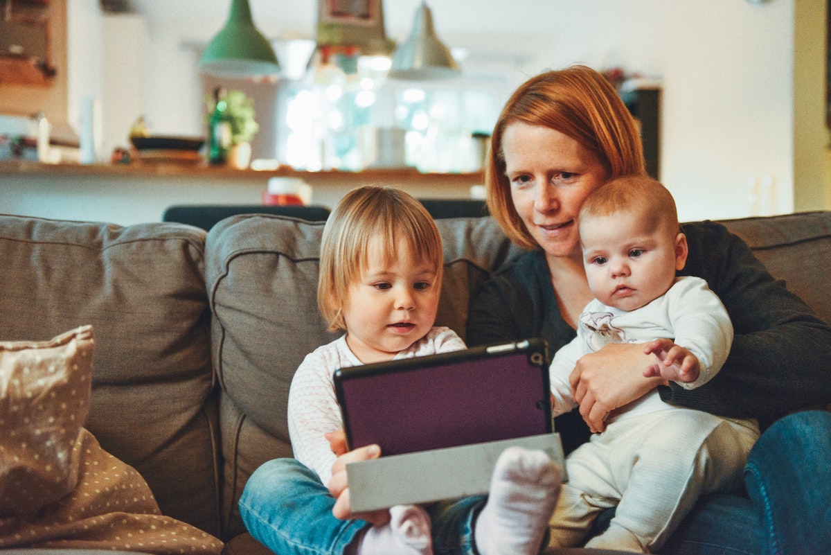 woman on couch looking at an tablet computer and holding two babies